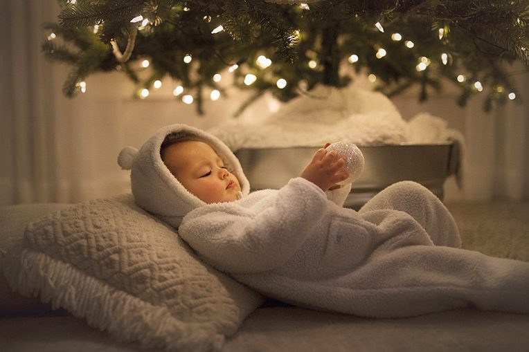 Baby in white bear onesie holds bauble under lit Christmas tree
iola joy issue Day of perfect being
